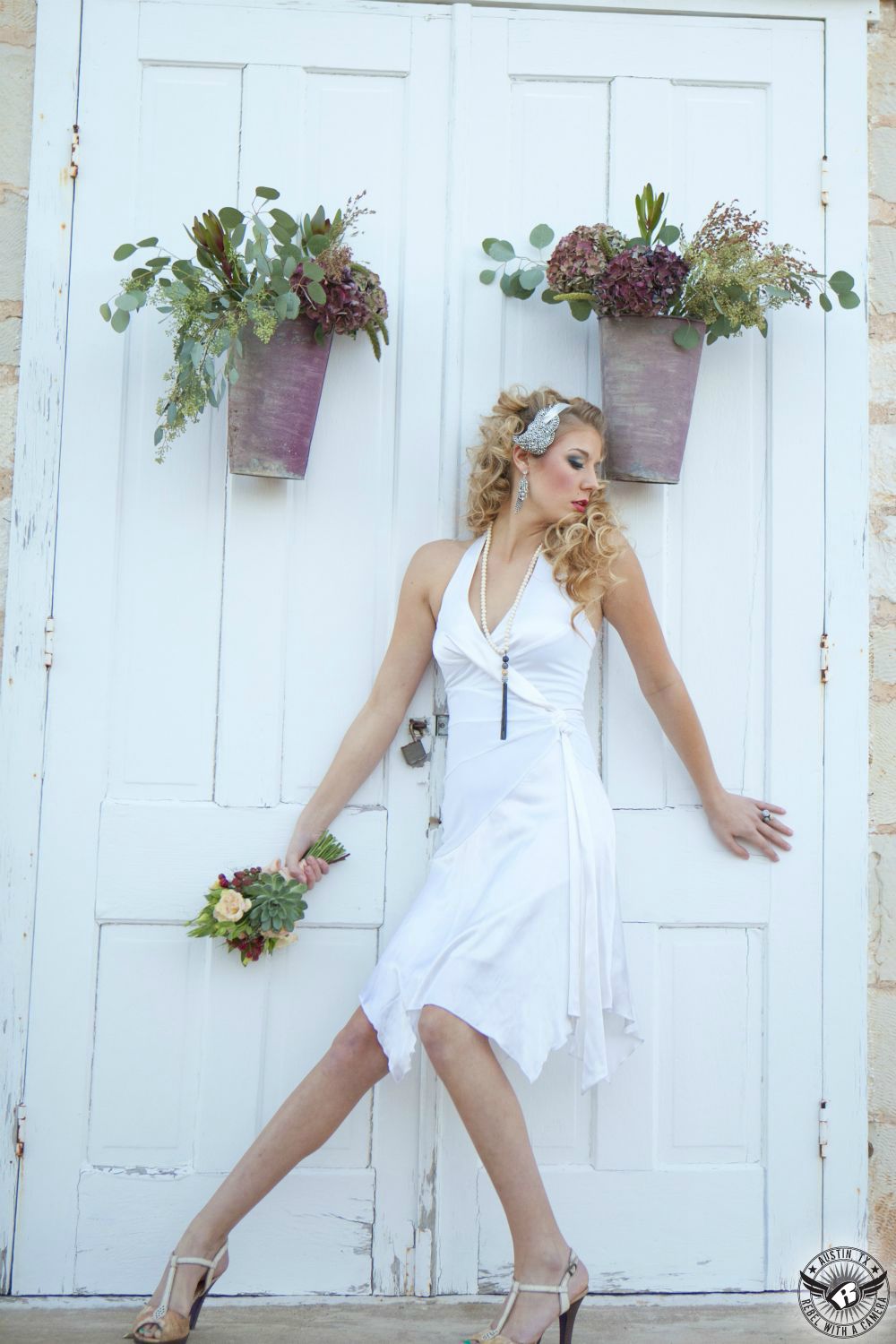 sexy blond female model with flowing white knee length 50 dress with low cut straps with blingy  vintage hairpiece leaning on a white antique double doors with hanging flower pots